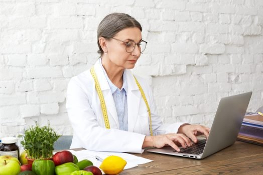 Focused middle aged woman nutritionist studying public health scientific research on food to treat nutritional problems, sitting at desk with various vegetables, keyboarding on portable computer